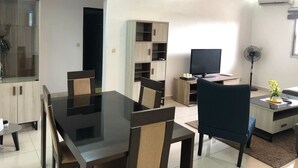 A modern living room with a glass-top dining table, dark chairs, a flat-screen TV, a standing fan, and built-in shelving. - Appartement Dayer (Abidjan)