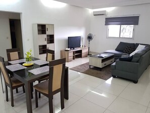 A modern living room with a dining area, a sofa, a TV, and a fan. - Appartement Dayer (Abidjan)