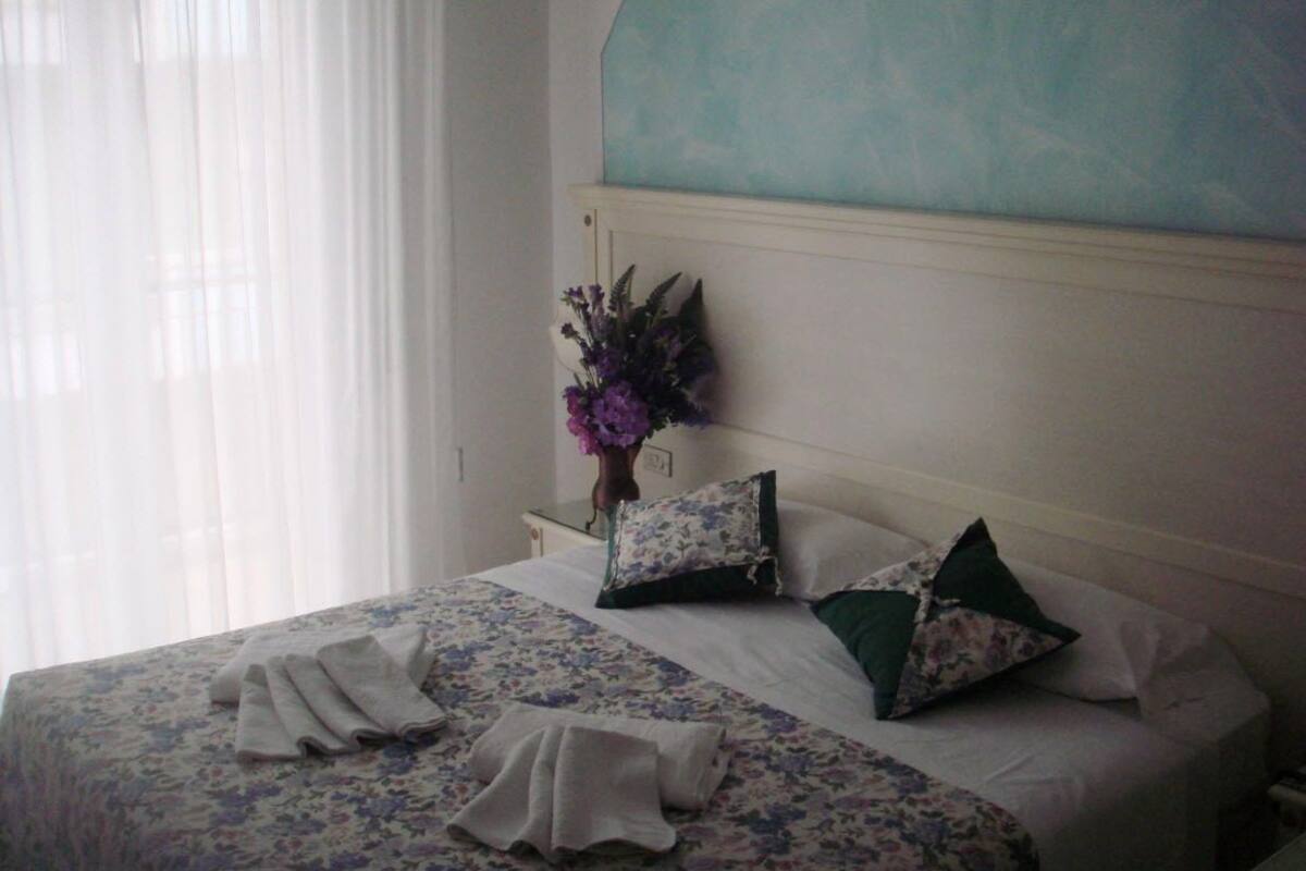 Un lit bien fait, avec une literie à motifs floraux, un vase rempli de fleurs violettes et un mur de couleur turquoise.