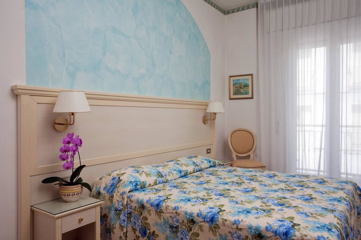 Une chambre à coucher avec une couvre-lit à motifs floraux, une table de chevet avec une lampe, une orchidée en pot et une chaise.