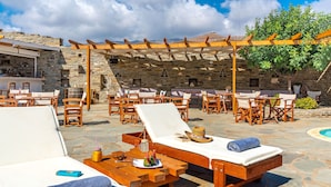 A sunlit outdoor seating area with lounge chairs and tables, stone walls, and a pergola providing shade. - Villa Sofia (Andros)