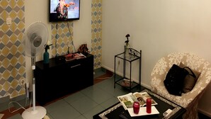 A living room with a TV mounted on the wall, a standing fan, a black entertainment unit, a magazine on a coffee table, and a patterned wall. - Résidence Bimbois (Abidjan)