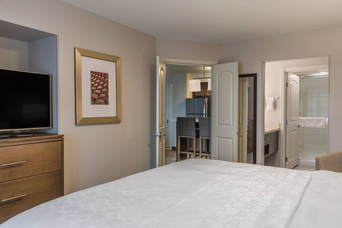 Ein Hotelzimmer mit Bett, Fernseher, einem gerahmten Bild und einer kleinen Küche im Hintergrund.