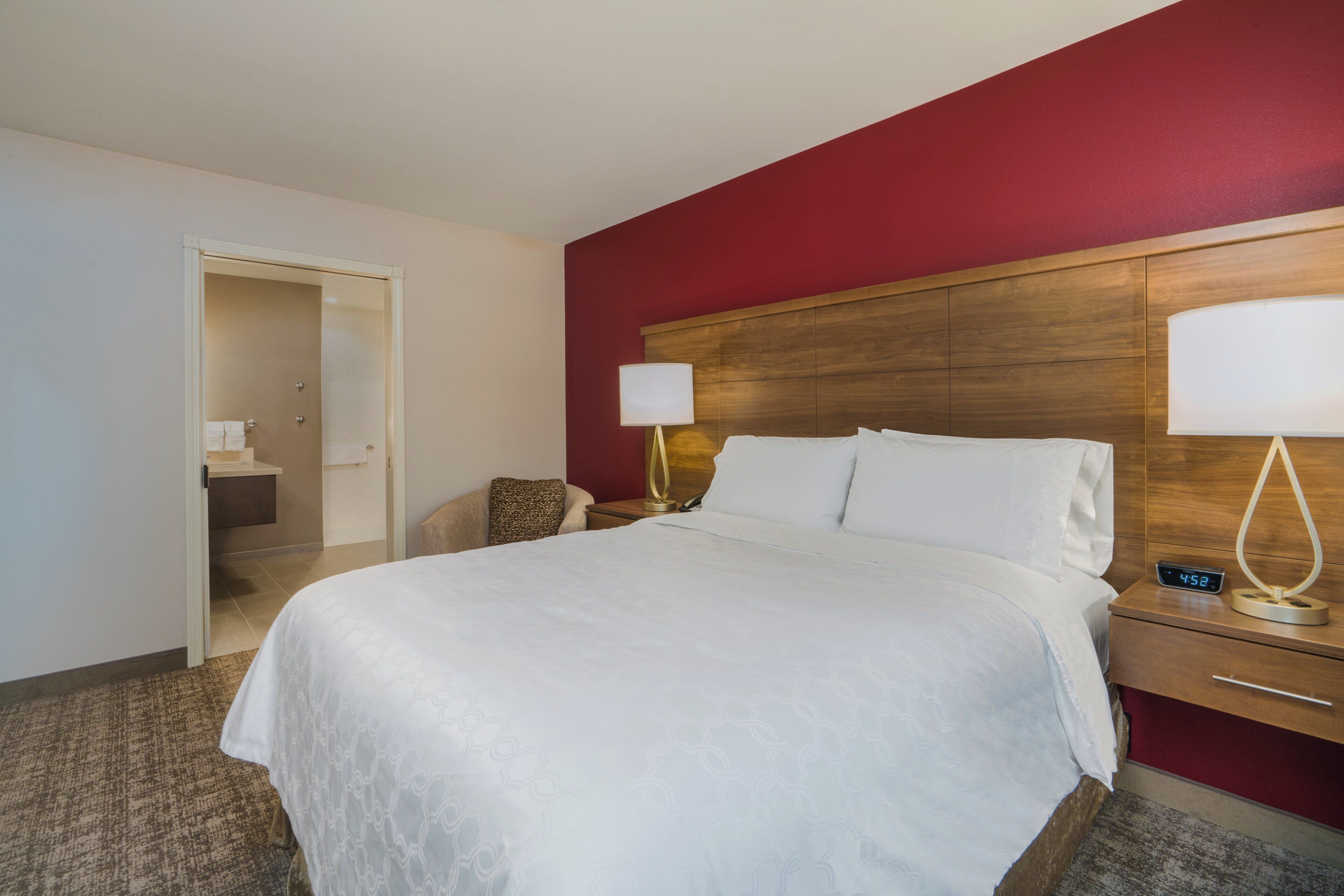 Habitación de hotel con una cama, una mesita de noche con lámpara, un reloj digital y una puerta que da al baño.