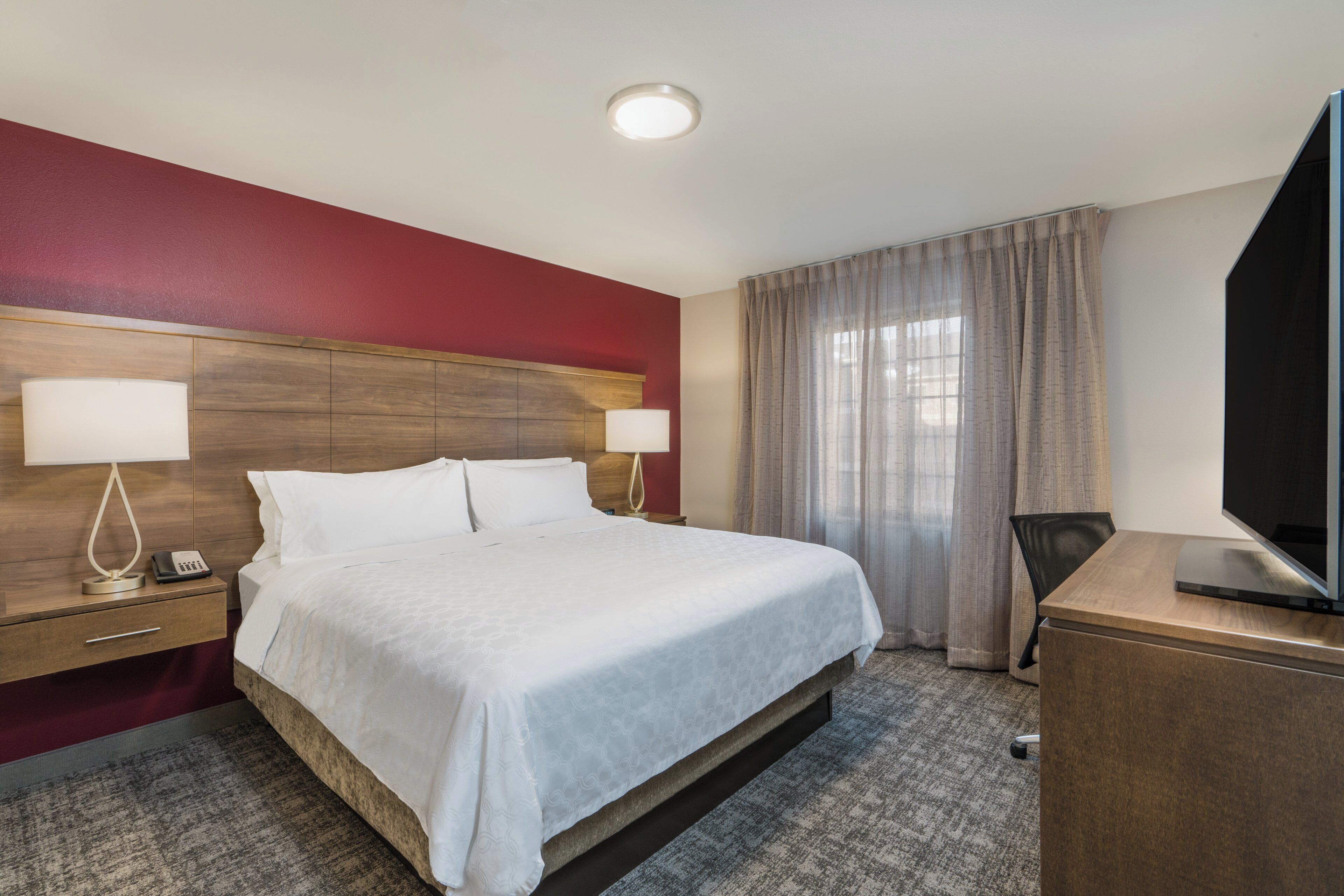 A hotel room with a bed, bedside tables, a desk, a television, and a window with curtains.