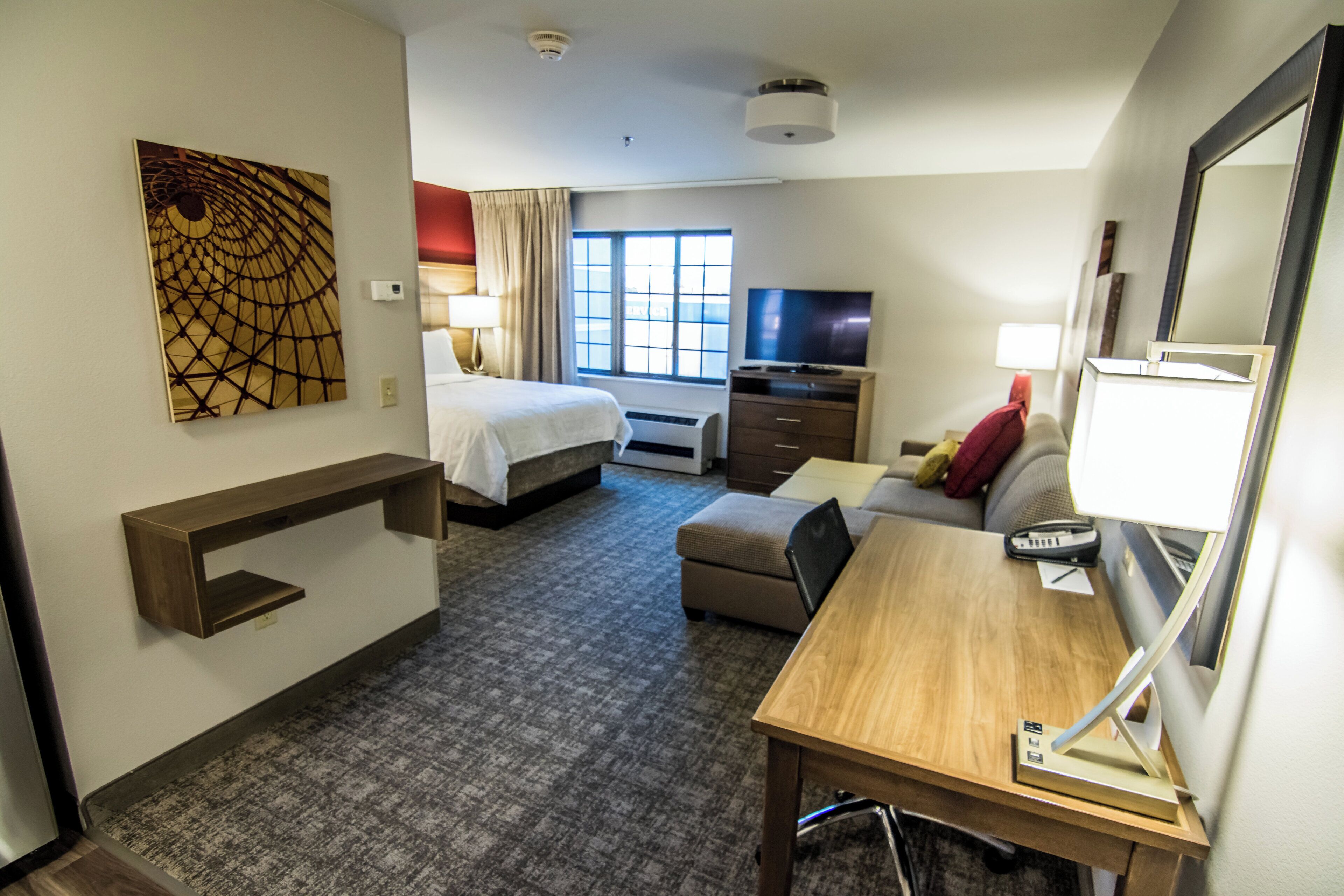 Habitación de hotel con cama, escritorio, televisor, espejo y un cuadro en la pared.