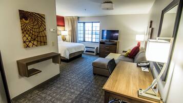 Habitación de hotel con cama, escritorio, televisor, espejo y un cuadro en la pared.