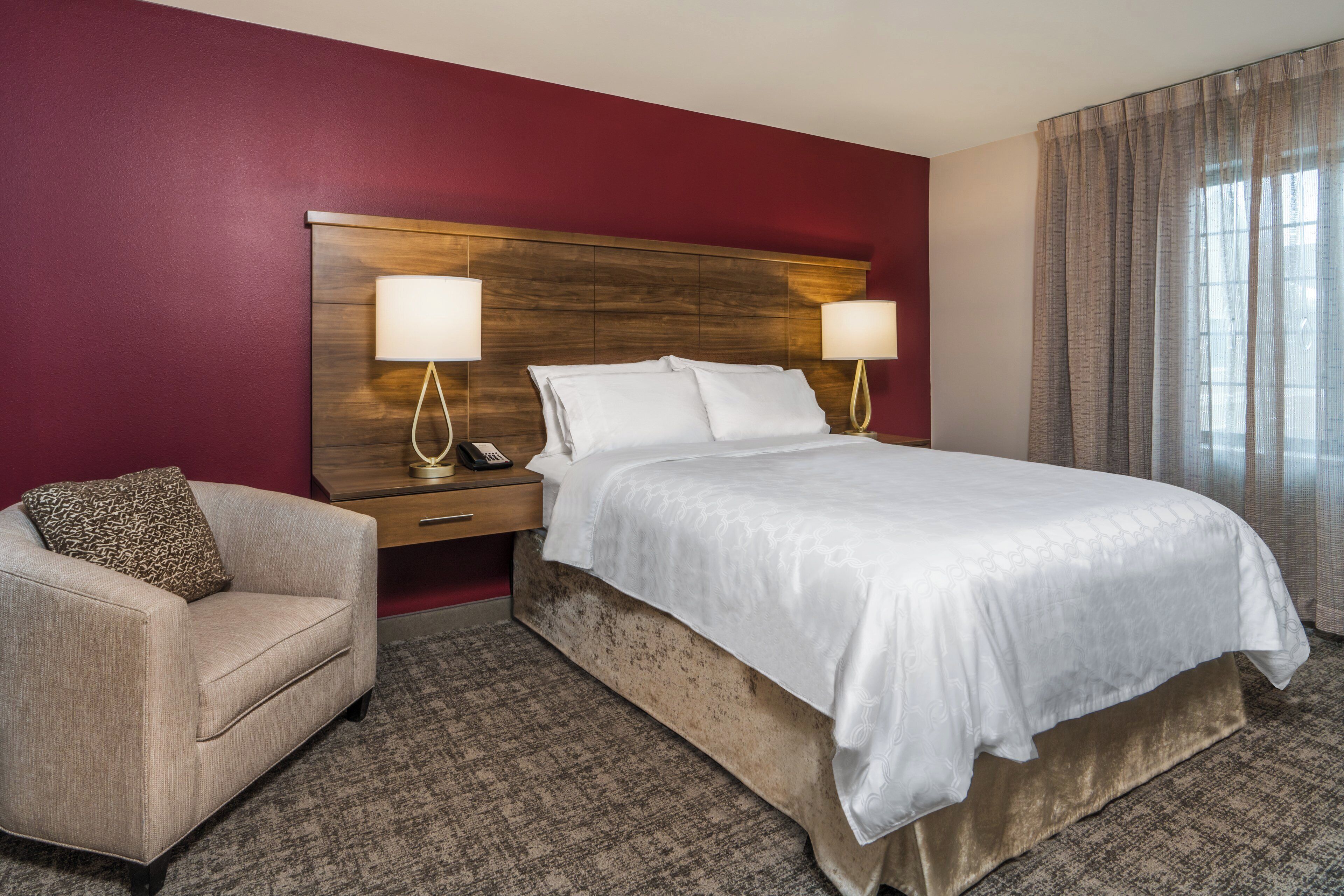 Habitació d'hotel amb un llit gran, dues làmpades de nit, una taula de nit, una cadira i una paret vermella decorativa.