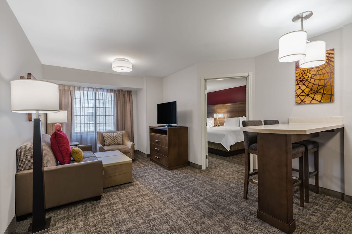 Ein Hotelzimmer mit einer Couch, einem Fernseher, einem Schreibtisch mit Stühlen, einem Bett und einem Bild an der Wand.