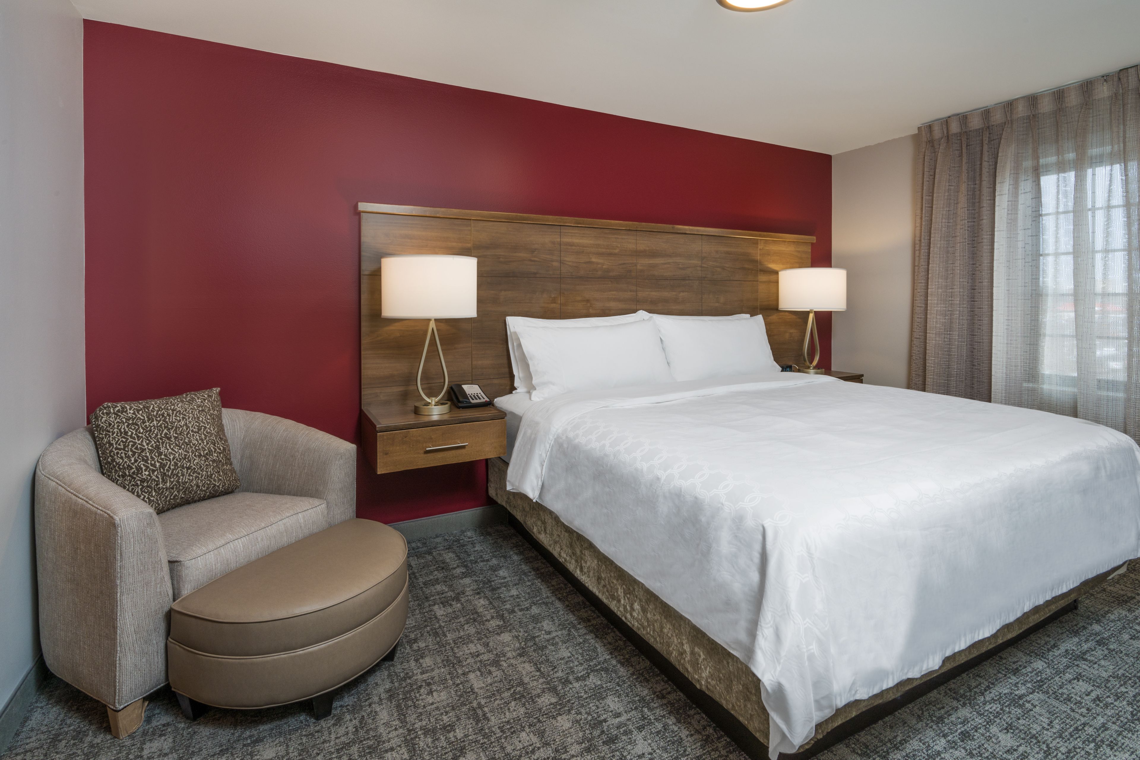 A hotel room with a large bed, two bedside lamps, a wooden headboard, a beige armchair, and a small table with a telephone.