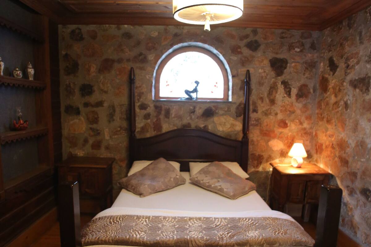Ein Bett mit Holzrahmen und Kopfteil, eine Steinsäule, ein kleines Fenster und ein Nachttisch mit Lampe.