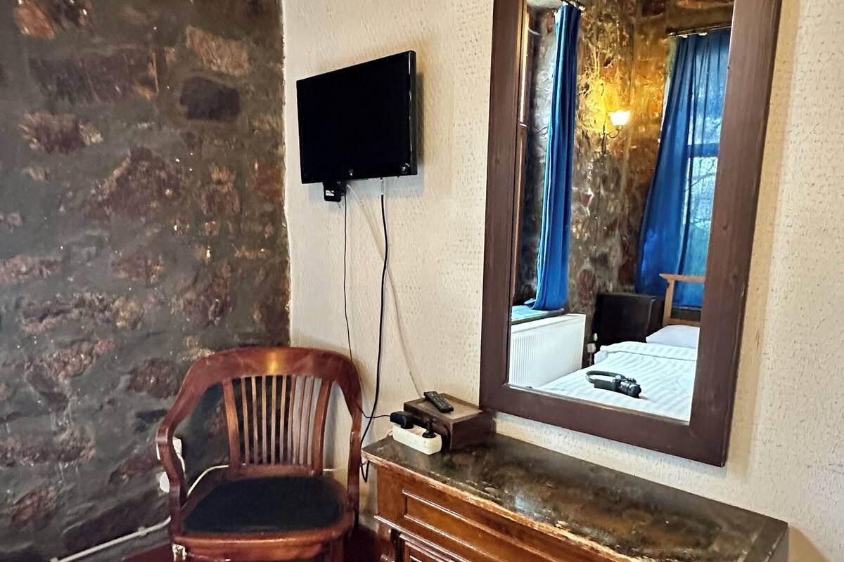 Ein Zimmer mit einer Steinsäule, einem Holzstuhl, einem Holzkonsolentisch, einem an der Wand befestigten Fernseher und einem Spiegel, der ein Bett mit blauen Vorhängen reflektiert.