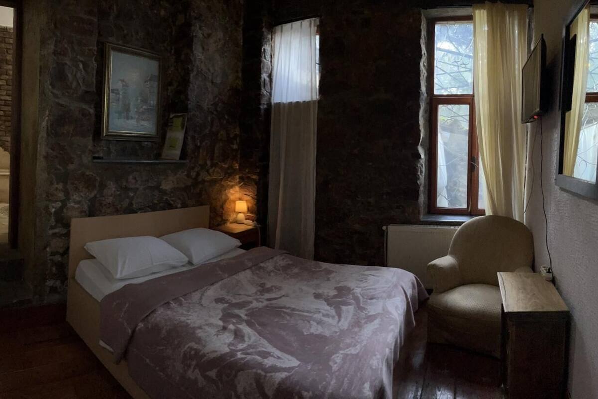 Ein Schlafzimmer mit Bett, Sessel, Nachttisch, Fenster mit Vorhängen, einem Bild an der Wand und einer Ziegelwand.