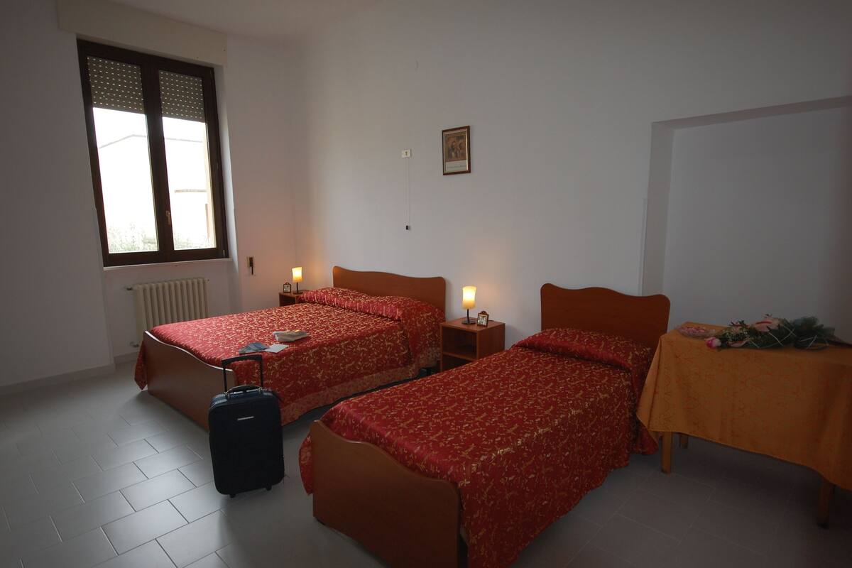 Habitación de hotel con dos camas, una mesa con un arreglo floral, una maleta y una ventana con persianas.