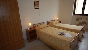 Une chambre avec deux lits, une armoire, une table de chevet avec une lampe, un tableau encadré au mur et une fenêtre donnant sur un paysage.