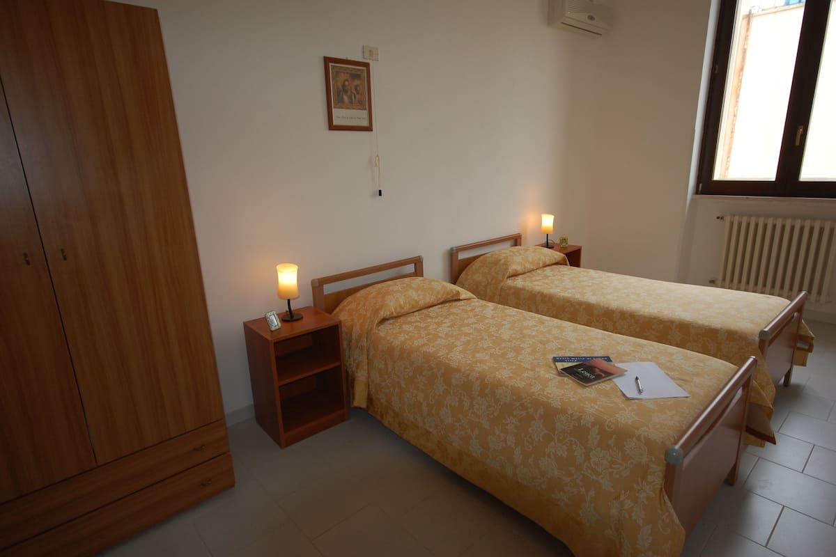 Un dormitorio con dos camas, un armario, una mesita de noche con lámpara, un cuadro en la pared y una ventana con vistas.