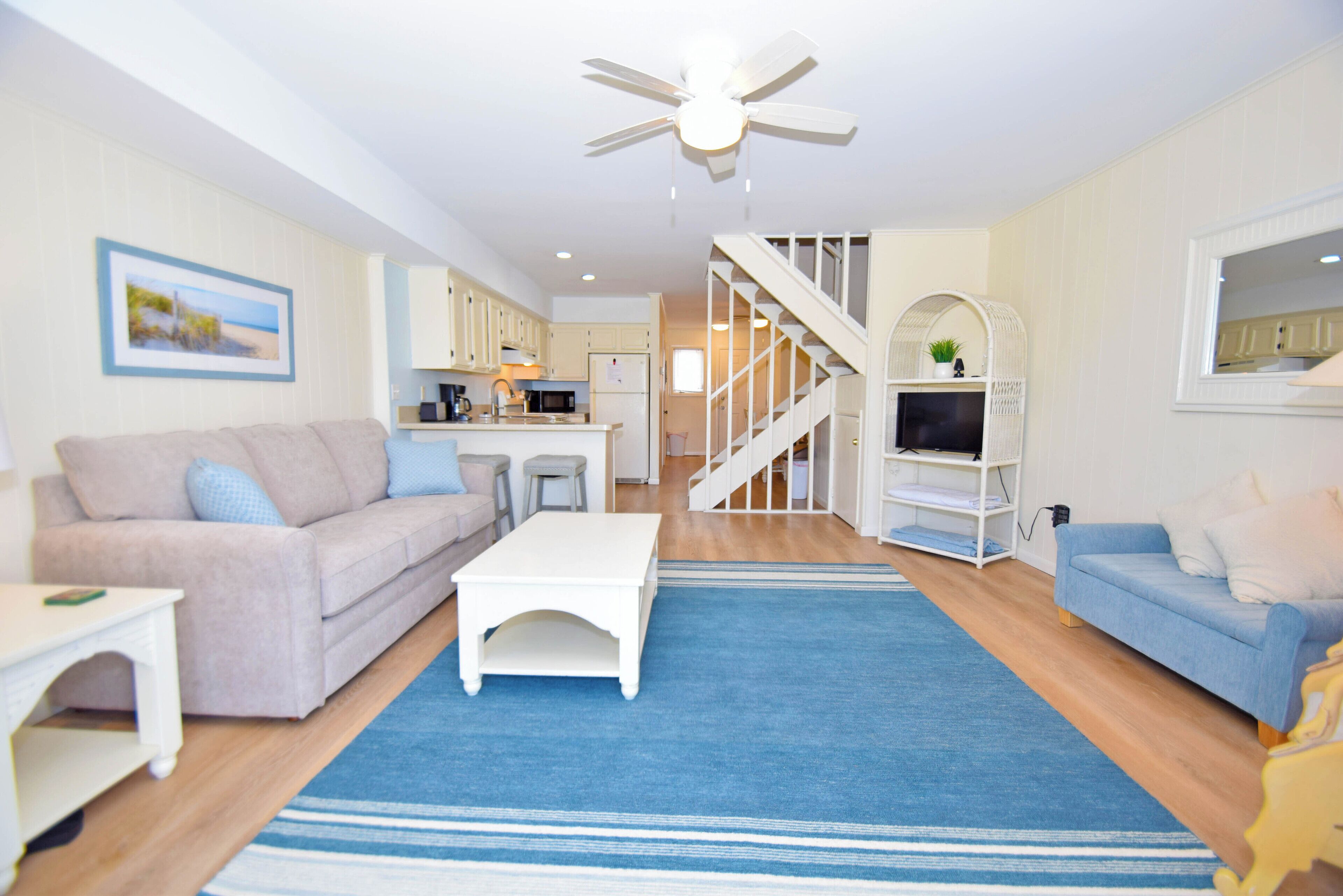 A spacious living room with a blue rug, a white coffee table, a beige sofa, and a blue sofa. A staircase leads to an upper level with a TV and a white shelf.