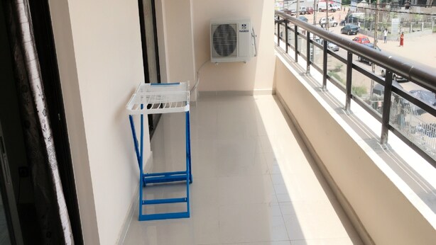 A balcony with a white air conditioning unit, a blue metal shelf, and a view of a busy street with buildings and cars.