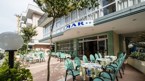 An outdoor seating area with tables and chairs, a building with a sign reading "MAR", and a tree in the foreground. - Hotel Belmar (Rimini)