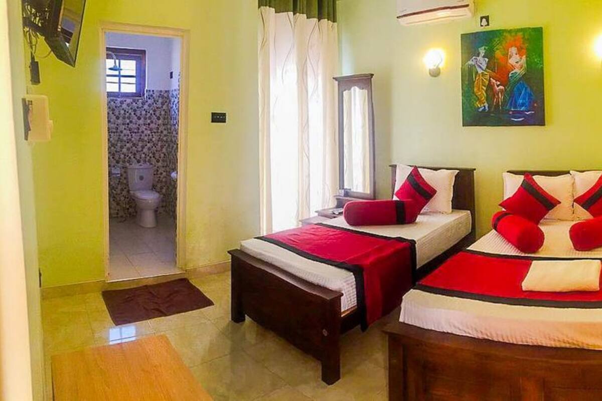 A room with two beds, a wooden bench, a window with curtains, a painting on the wall, and a door leading to a bathroom.