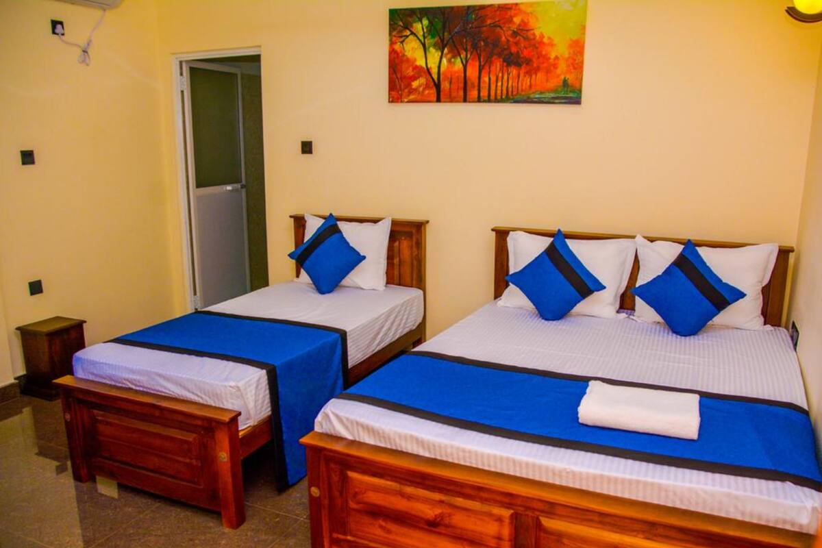 Two single beds with blue bedding and white pillows, a wooden bench, a small bedside table, and a painting of trees on the wall.