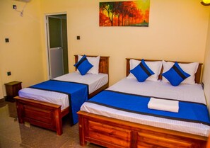 Two single beds with blue bedding and white pillows, a wooden bench, a small bedside table, and a painting of trees on the wall. - Marathona Tourist Resort - Anuradhapura (Anuradhapura)