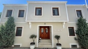 A two-story building with a beige facade, a wooden front door, and the name "AIKA" above the entrance. There are two potted plants on either side of the door. - Aika Hotel Alaybey (Bozcaada)
