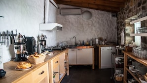 A rustic kitchen with a stone wall, wooden beams, a built-in stove, and a variety of kitchen utensils and appliances. - Aika Hotel Alaybey (Bozcaada)