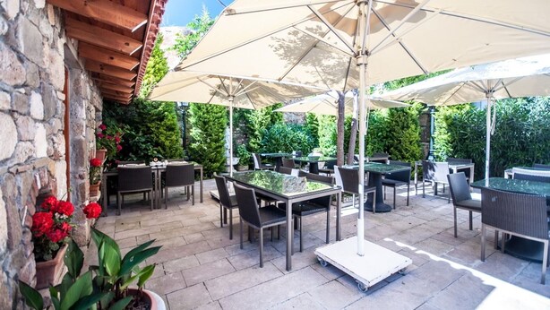 An outdoor patio with tables and chairs, surrounded by greenery and stone walls. - Aika Hotel Alaybey (Bozcaada)