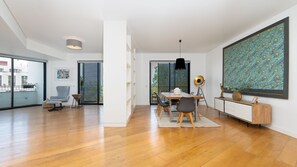 A spacious living area with a dining table, chairs, a sofa, and a large window with blinds.