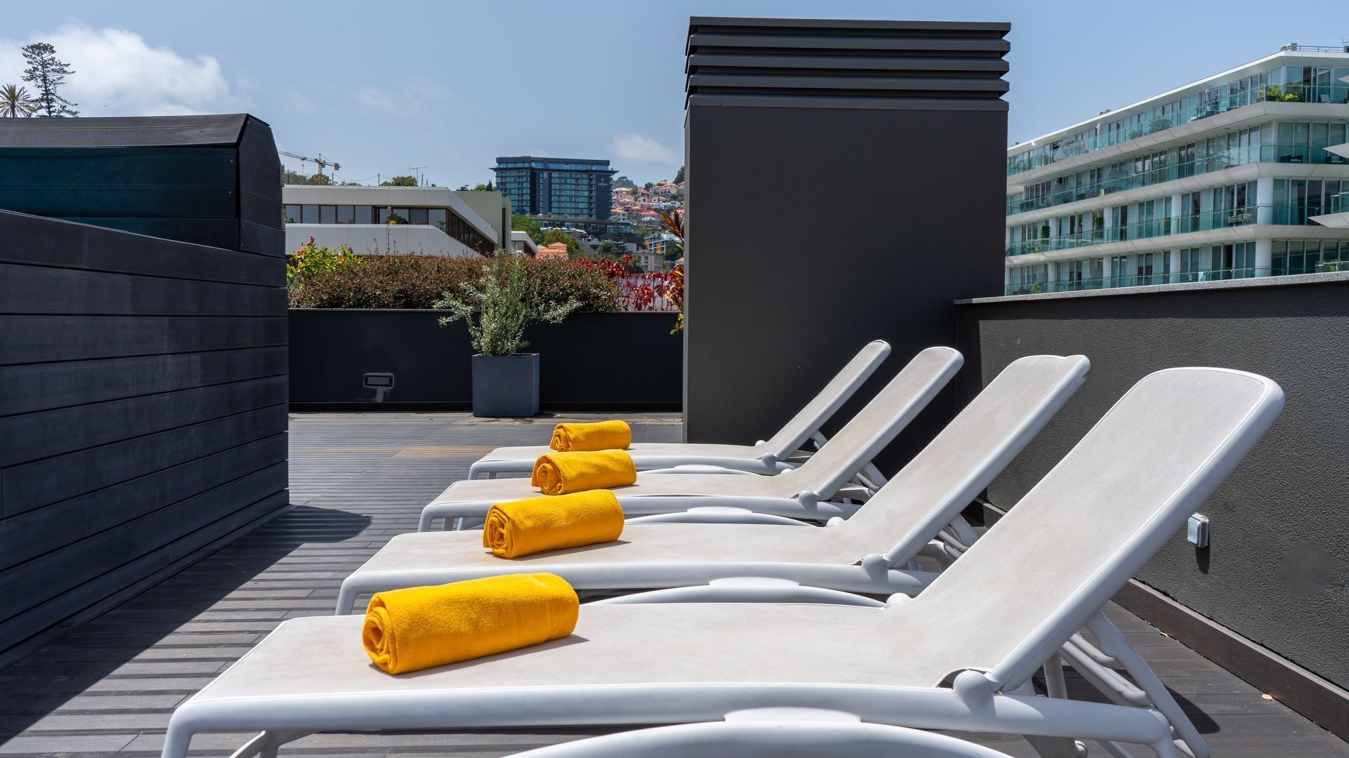 Eine Dachterrasse mit weißen Liegestühlen und gelben Handtüchern, im Hintergrund ein modernes Gebäude und ein klarer Himmel.