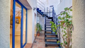 Un étroit couloir extérieur avec une porte à cadre bleu, un escalier en métal et des plantes en pot.