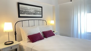 Una cama bien hecha con almohadas blancas y moradas, dos lámparas de noche y una fotografía en blanco y negro enmarcada en la pared.