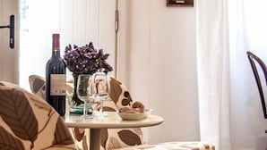 Un comedor con una mesa preparada para dos personas, que incluye una botella de vino, copas y un jarrón de flores. Hay dos cuadros en la pared y una silla con un cojín estampado con motivos de hojas.