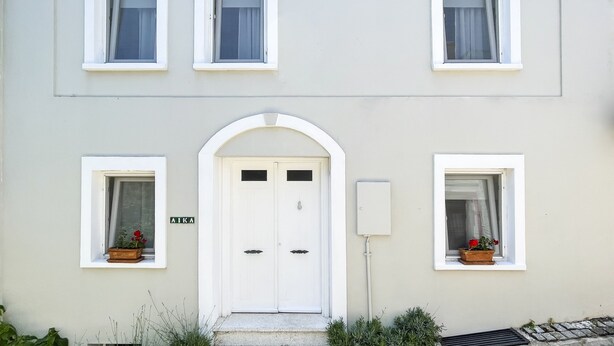 Un edificio con fachada blanca, una puerta doble central y cuatro ventanas con jardineras.