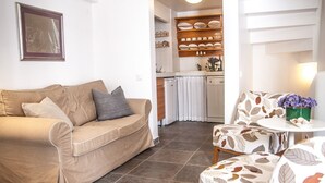 Una sala de estar con un sofá color crema, un sillón estampado y una mesita con un jarrón de flores. Se ve una cocina al fondo, con un refrigerador blanco y una repisa con vajilla.