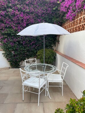 Une table ronde en verre, entourée de chaises en métal blanc et surmontée d’un parasol blanc, est placée sur une terrasse agrémentée d’un mur décoratif et de fleurs violettes en arrière-plan.