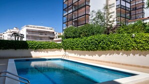Una piscina rectangular con una escalera de metal, rodeada por un seto bajo y un muro blanco.