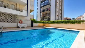 Une piscine à l’eau d’un bleu limpide, entourée d’un bâtiment blanc doté de balcons et d’une haie.