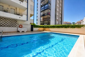 A swimming pool with a clear blue water surface, surrounded by a white building with balconies and a hedge. - The Rose by Hello Homes Sitges (Sitges)