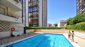 A swimming pool with a clear blue water surface, surrounded by a tiled deck and a low hedge. - The Rose by Hello Homes Sitges (Sitges)