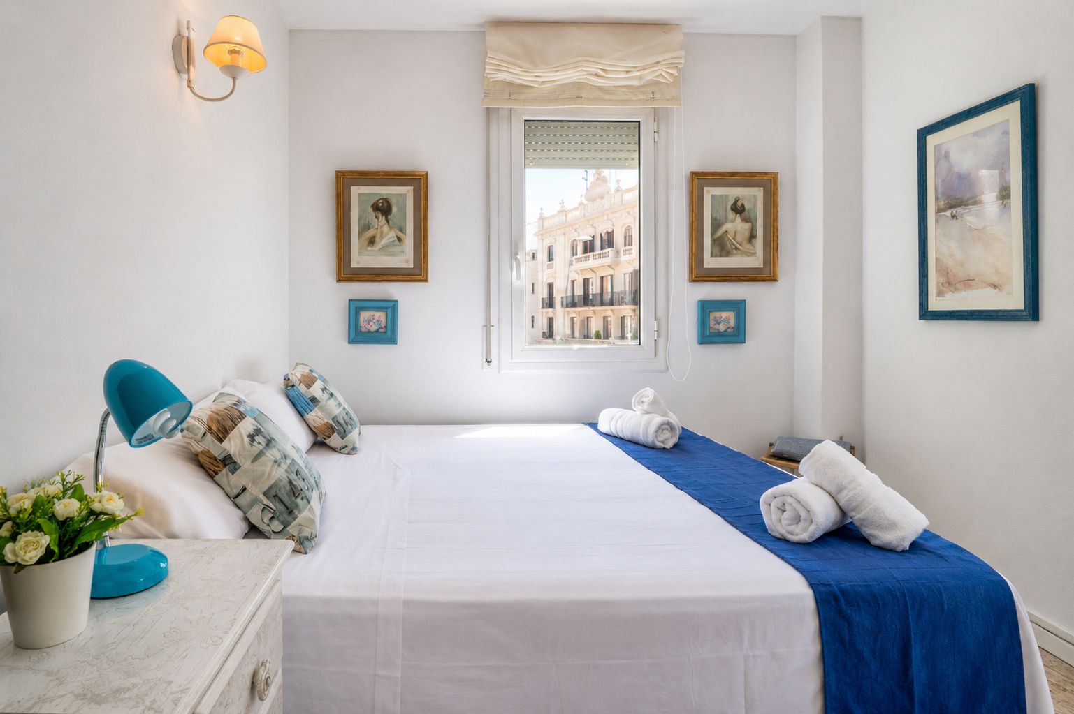 Una camera d'albergo con un letto, una scrivania con lampada, una finestra vista edifici e opere d'arte alle pareti.
