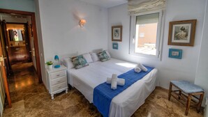 Un dormitorio con cama, mesita de noche, lámpara y un taburete pequeño.