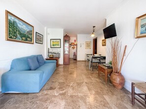 A living room with a blue sofa, a dining table with chairs, a TV mounted on the wall, and a fireplace. - The Rose by Hello Homes Sitges (Sitges)