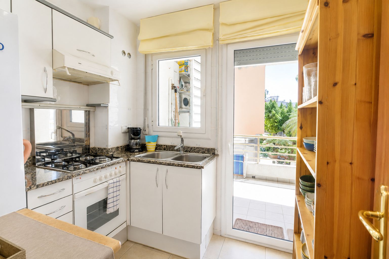 Une cuisine compacte avec des meubles blancs, une cuisinière à gaz, un évier et une fenêtre donnant sur un balcon agrémenté de plantes en pot.