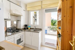 A compact kitchen with white cabinets, a gas stove, a sink, and a window overlooking a balcony with potted plants. - The Rose by Hello Homes Sitges (Sitges)