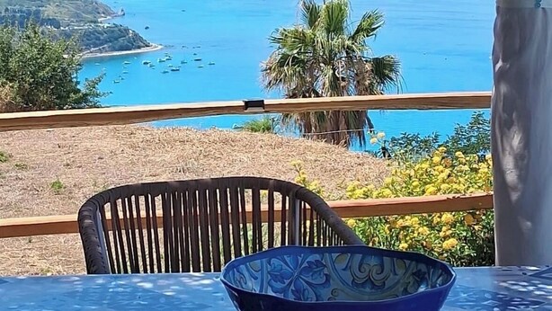 A table with a patterned tablecloth, a blue bowl, and a chair, overlooking a coastal view with palm trees and the sea. - Sea view villa with pool near Tropea (Zambrone)