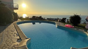 A swimming pool with a curved edge, a stone-tiled deck, lounge chairs, and a view of the sea at sunset. - Sea view villa with pool near Tropea (Zambrone)