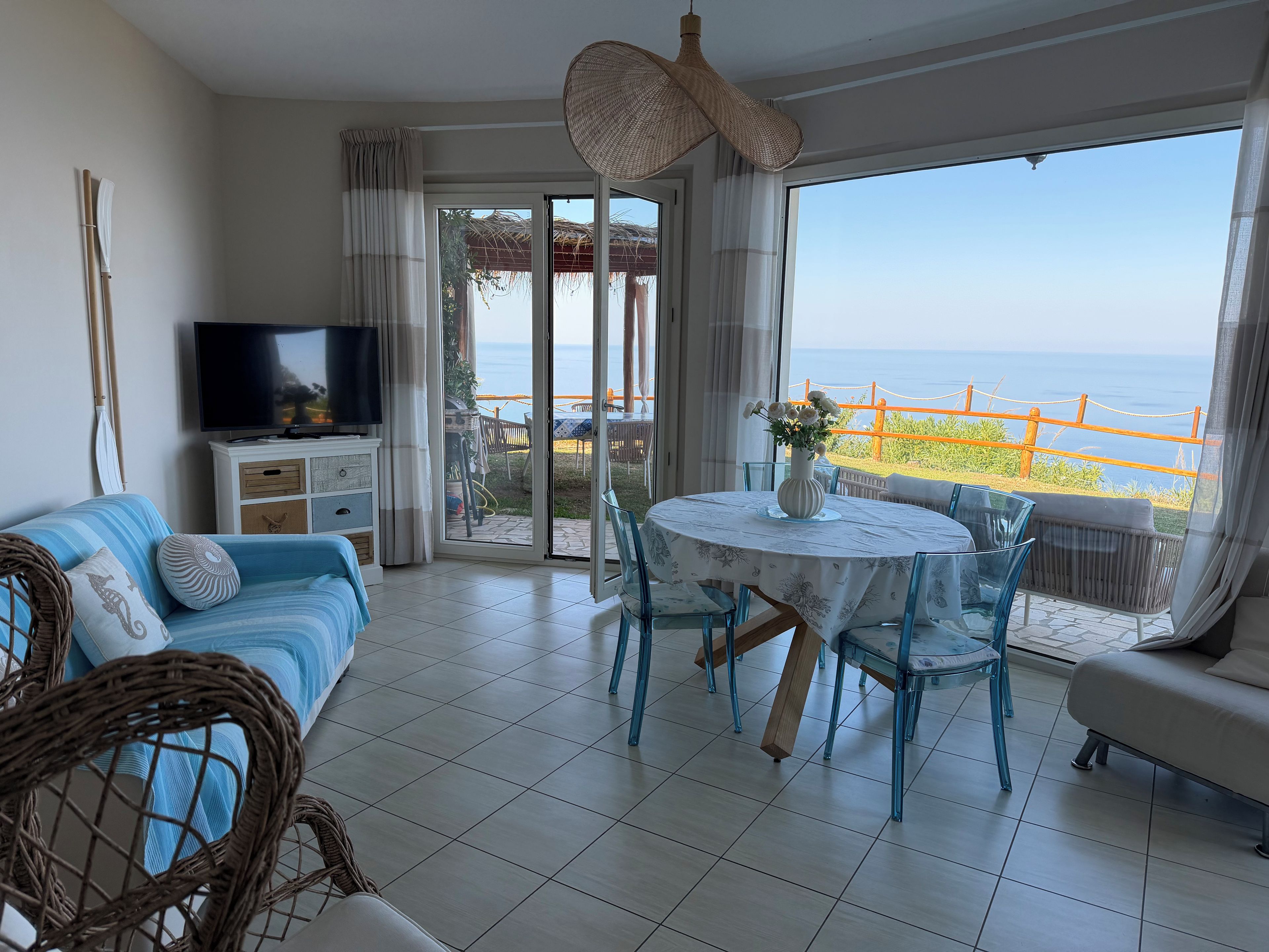 Sala de estar con sofá, televisor y una mesa con sillas. Hay una puerta corrediza de vidrio que da a un balcón con vista al mar.