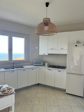 A modern kitchen with white cabinets, a built-in gas stove, a Samsung refrigerator, a window with a view, and a wicker pendant light.